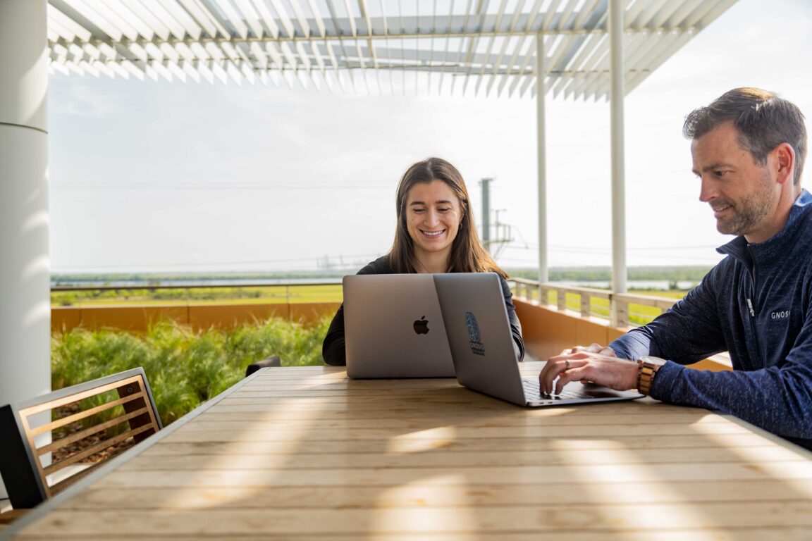 A male and female sit smiling and working on laptops on a patio