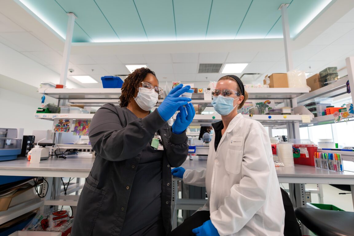 A black and a white female employees in lab gear inspect a medicine jar