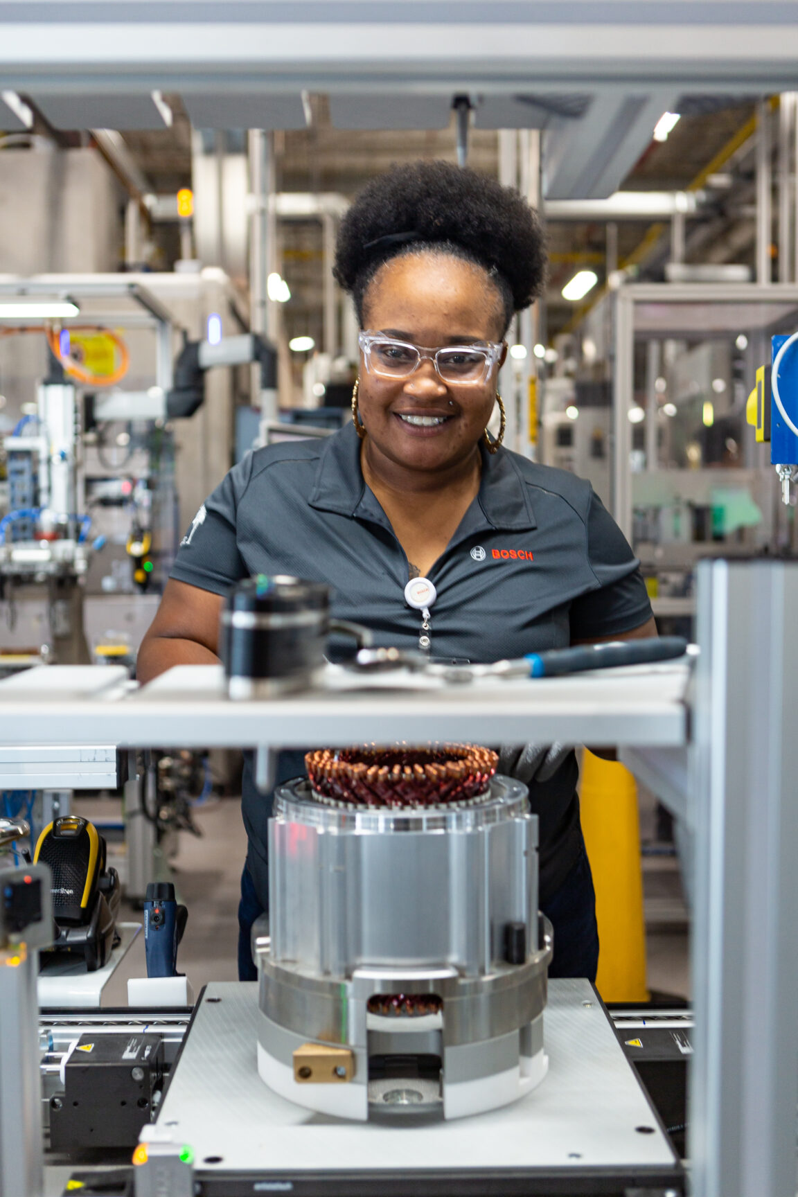 Black woman wearing safety glasses stands in front of heavy machinery, smiling