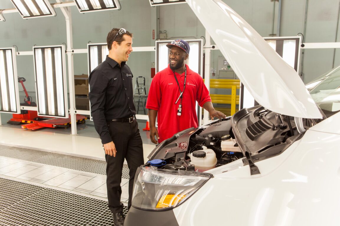 A black and a white male employee stand in front of an open car hood